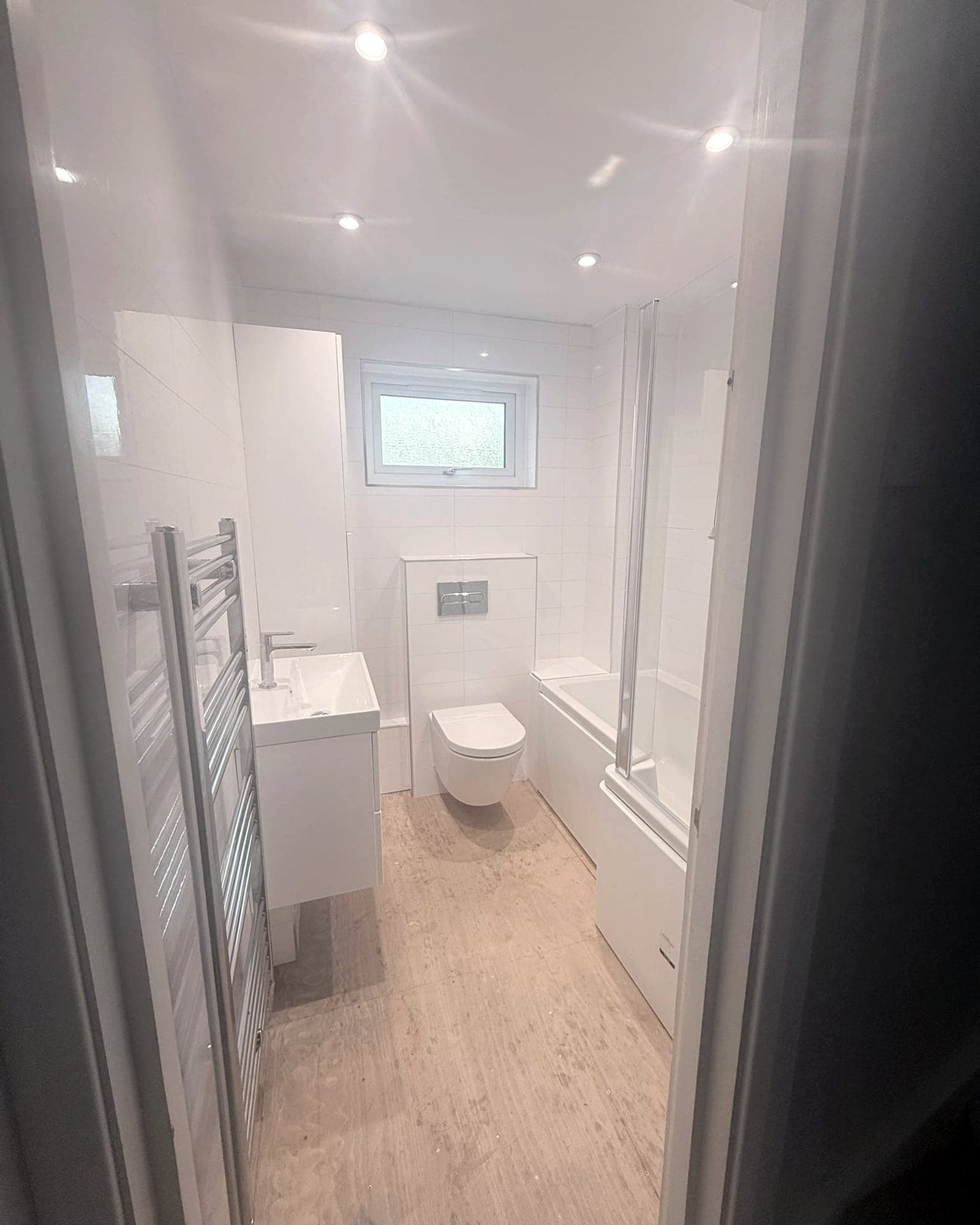 A finished bathroom seen through the doorway: white tiles, a chrome towel radiator, a wall-hung toilet and basin
