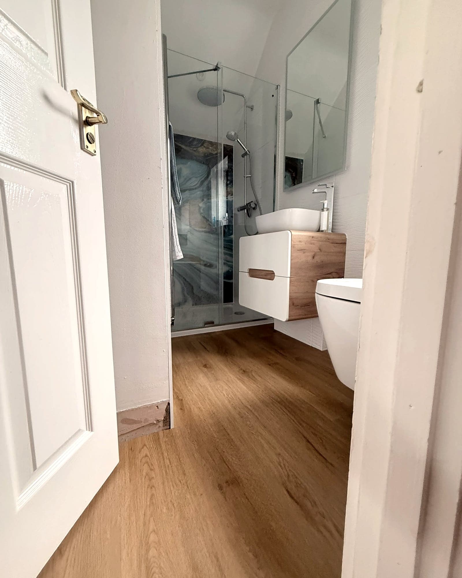 Looking through an open door into an en suite with a wood-effect floor, a glass shower and a basin unit